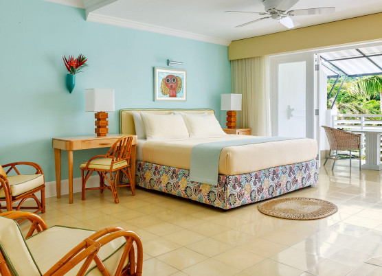 bedroom with light blue walls, light yellow linens and chairs, blue draped quilt, and balcony overlooking the ocean