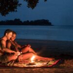 couple sitting on a blanket with a candle having drinks on the beach at night